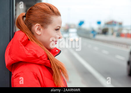 Die junge rothaarige Frau, die an der Bushaltestelle sie das Taxi draußen erwartet. Junge rothaarige Frau in rotem Mantel stehen draußen, im Freien in autu Stockfoto