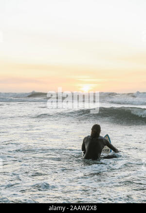 Ansicht der Rückseite des Surfer mit Surfboard sitzen auf Surfbrett im Meer bei Sonnenuntergang Stockfoto