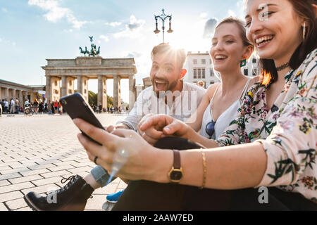 Drei lachen Freunde sitzen in der Nähe von Brandenburger Tor am Smartphone, Berlin, Deutschland Stockfoto
