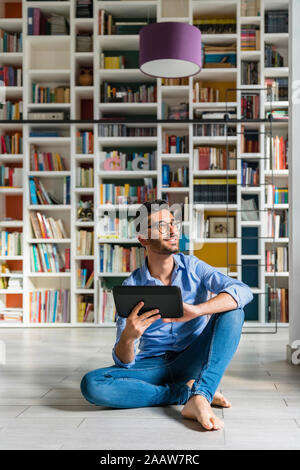 Portrait von barfüßigen Jungen Mann sitzt vor bücherregale auf dem Boden mit digitalen Tablet Stockfoto