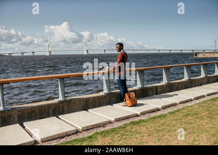Junge Mann sitzt auf Geländer am Meer, Hamburger essen, trinken Kaffee Stockfoto