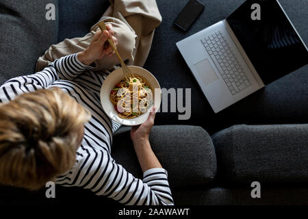 Reife Frau mit Laptop Essen hausgemachte Pasta auf der Couch zu Hause Stockfoto