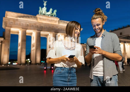 Glückliches Paar mit Smartphones am Brandenburger Tor bei Nacht, Berlin, Deutschland Stockfoto