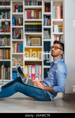 Portrait von nachdenklicher junger Mann sitzt vor bücherregale auf dem Boden mit Laptop Stockfoto
