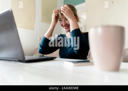 Lachende Frau am Tisch zu Hause am Laptop suchen Stockfoto