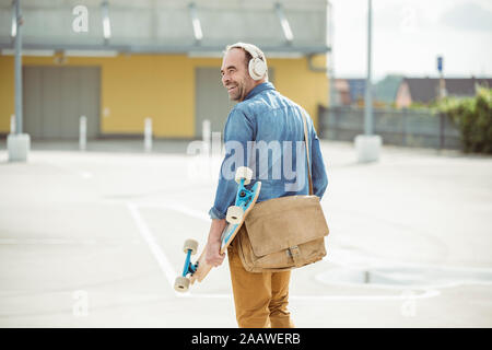 Gerne casual reifer Mann mit Kopfhörern und Skateboard zu Fuß auf dem Parkdeck Stockfoto