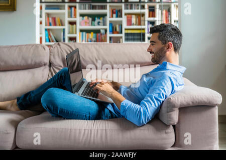 Lächelnden jungen Mann auf der Couch liegen zu Hause mit Laptop Stockfoto