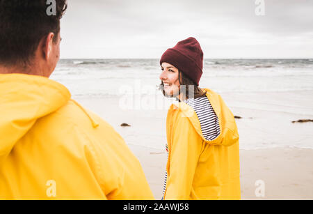 Junge Frau tragen gelbe Regenjacken und zu Fuß am Strand entlang, Bretagne, Frankreich Stockfoto