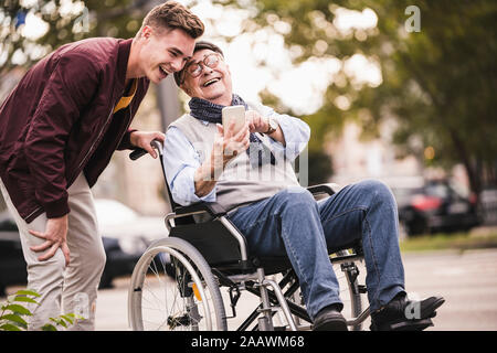 Lachend älterer Mann im Rollstuhl und seines erwachsenen Enkel zusammen am Smartphone Spaß suchen