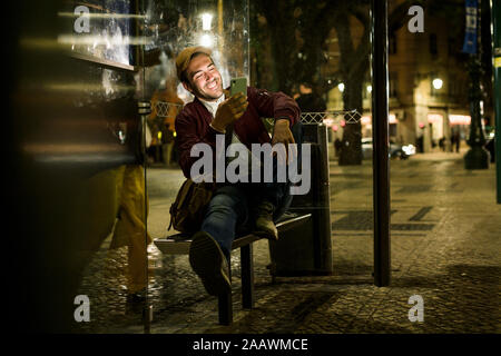 Portrait von lachenden Jungen Mann an der Bushaltestelle in der Nacht mit Smartphone sitzen, Lissabon, Portugal Stockfoto