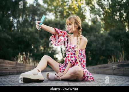 Portrait von lächelnden jungen Frau sitzt auf der Promenade im Sommer unter selfie mit Smartphone Stockfoto