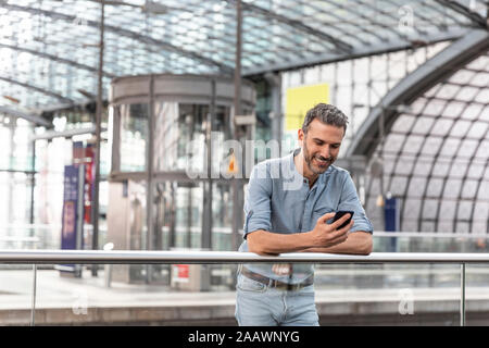 Lächelnder Mann am Bahnhof, der auf das Smartphone schaut, Berlin, Deutschland Stockfoto