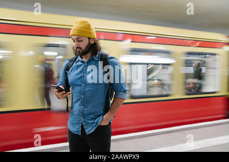 Bärtiger Mann mit Rucksack auf Smartphone suchen und gleichzeitig Plattform, Berlin, Deutschland Stockfoto