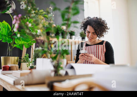 Lächelnde junge Frau mit Handy in einem kleinen Laden mit Pflanzen Stockfoto