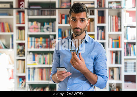 Portrait von skeptischen jungen Mann mit Smartphone stand vor der Bücherregale Stockfoto
