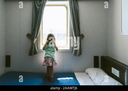 Zurück Blick auf kleine Mädchen stehen auf Bett zum Meer Blick aus dem Fenster des Schiffs Kabine Stockfoto