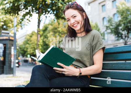 Portrait von lächelnden jungen Frau sitzt auf der Bank mit einem Buch Stockfoto