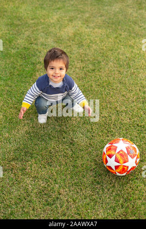 Portrait von lächelnden kleinen Jungen mit einem Ball zusammengekauert auf Rasen Stockfoto