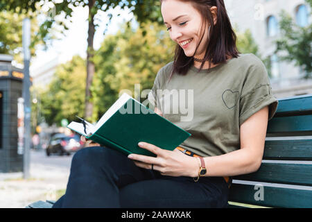 Portrait von lächelnden jungen Frau sitzt auf der Bank ein Buch lesen Stockfoto