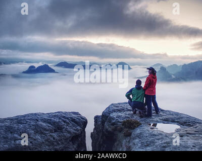 Nacht Foto einer Frau zeigt magische Blitze Laterne zu ihrem Mann. Frau sitzt auf einem Felsen und glänzt der Finsternis Misty. Erste Sonnenstrahlen erscheinen in der Clou Stockfoto