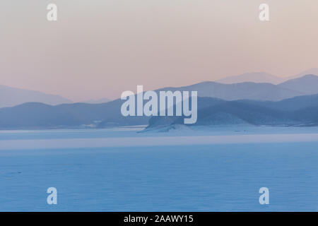 Winterlandschaft von Sonnenuntergang am See Songhua gegen Schnee Berge und zugefrorenen See mit Nebel auf dem See Oberfläche, Provinz Jilin, China Stockfoto