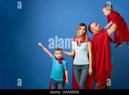 Glückliche Mama und Papa, Kinder Superhelden in roten Mänteln auf leeren blauen Hintergrund im Studio Stockfoto