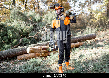 In voller Länge Porträt eines professionellen lumberman in schützende Arbeitskleidung mit einer Kettensäge und Baumstämmen, der während der Arbeit auf die Protokollierung im Pinienwald Stockfoto
