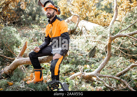 Porträt eines professionellen lumberman in schützende Arbeitskleidung sitzen auf den gefällten Baum beim Ausruhen nach der harten Arbeit im Wald Stockfoto