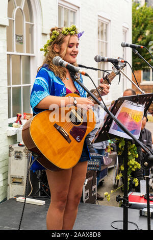 Maisy Beth, junge blonde Volkssänger, Gesang während der Wiedergabe akustischer Gitarre in die Straße an der Faversham Hop Festival. Trägt Girlande von Hopfen. Stockfoto