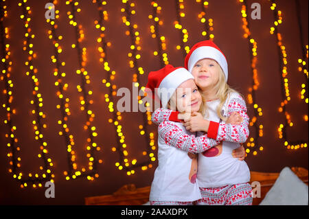 Zwei süße kleine Schwestern in santa Caps sind umarmt, während im Zimmer stehend vor dem Hintergrund der hellen festliche Lichter. Warten auf den Urlaub Stockfoto