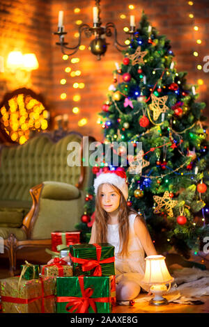 Ein nettes Mädchen in einem weißen Kleid und Santa Claus hat ist das Sitzen auf dem Boden und einen großen Haufen von Geschenken. Vor dem Hintergrund der Festliche Baum. Christm Stockfoto