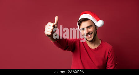 Junge stattlichen kaukasischen Kerl im roten Pullover und Nikolausmützen steht auf roten Hintergrund im Studio und zeigt den Daumen nach oben. Close up Portrait europäischen Kerl wi Stockfoto