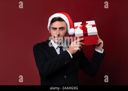 Junge stattlichen kaukasischen Typ in Anzug und Nikolausmützen steht auf weißem Hintergrund im Studio und Smilie hält in der Nähe des Ohrs roter Geschenkbox Portrait Stockfoto