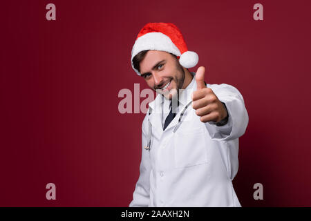 Jungen gutaussehenden Arzt in Weiß uniforme und Santa Claus hat im Studio auf rotem Hintergrund Lächeln und zeigt einen Daumen nach oben anmelden Porträt medizinischen s Stockfoto