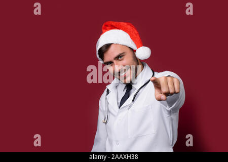 Jungen gutaussehenden Arzt in Weiß uniforme und Santa Claus hat im Studio auf rotem Hintergrund Lächeln und zeigt mit dem Finger in die Kamera Portrait medizinischen s Stockfoto