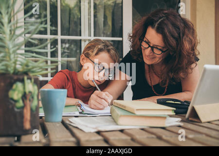 Müde Schüler Hausaufgaben zu Hause mit Schule Bücher, Zeitung und digitale Pad von seiner Mutter geholfen. Mom schreiben auf dem copybook Lehre seinen Sohn. Stockfoto