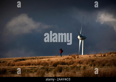 Walker auf dem Hügelkamm, der von Windturbinen mit dramatischem Himmel am späten Nachmittag in den South Wales Valleys in den Schatten gestellt wurde. Stockfoto