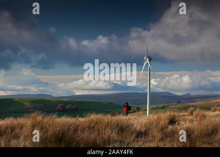 Walker auf dem Hügelkamm, der von Windturbinen mit dramatischem Himmel am späten Nachmittag in den South Wales Valleys in den Schatten gestellt wurde. Stockfoto