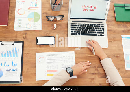 Ansicht von oben Hintergrund der unkenntlich Geschäftsfrau mit Laptop während der Arbeit am Schreibtisch aus Holz mit Dokumenten und Worte Marketingstrategie auf dem Bildschirm Platz kopieren Stockfoto