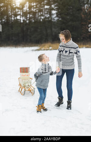 Hübsche, junge Frau mit ihrem Sohn junge Rollen Holz Schlitten mit vorhanden Boxen eingerichtet. Ein Kind geht mit seiner Mutter durch den Winterwald. Winter Stockfoto