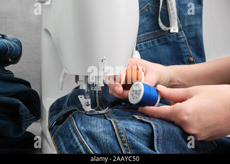 Nähen Jeans auf der Nähmaschine in der Werkstatt. Jeans reparieren. Die Frau entscheidet die Farbe der Nähfaden aus zwei Spulen. Stockfoto