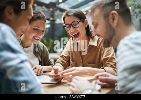 Gruppe von Jugendlichen am Bildschirm des Smartphones und fröhlich lachend beim gemeinsamen Mittagessen im Café genießen. Stockfoto