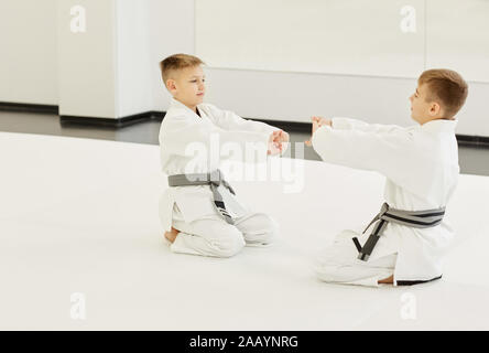 Zwei kleine Jungen im weissen Kimono auf dem Fußboden sitzen einander gegenüber und die Ausübung vor dem Training Stockfoto