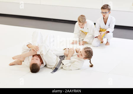 Mädchen kämpfen mit Jungen auf dem Boden, während andere Kinder jubeln für Sie während der Lektion im Karate in der Turnhalle Stockfoto