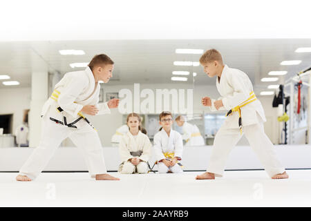 Zwei Jungen stehen einander gegenüber und lernen zu kämpfen, während andere Kinder sitzen auf dem Boden und Beobachten für Sie während der Lektion im Karate Stockfoto