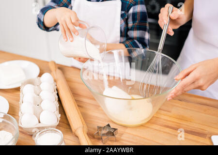 Kleines Kind Zucker in den Topf mit rohen Eiern beim Helfen seiner Mama mit Teig für hausgemachte Backwaren von Tisch in der Küche geschüttelt Stockfoto