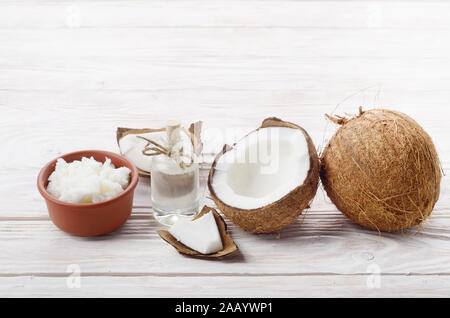 Hintergrund der Kokosnuss, Kokosnussschalen, Öl in Ton Schüssel und Glas auf weissem Holztisch Stockfoto