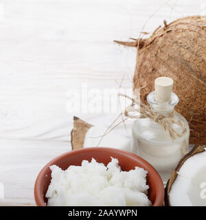 Hintergrund der Kokosnuss, Kokosnussschalen, Öl in Ton Schüssel und Glas auf weissem Holztisch Stockfoto