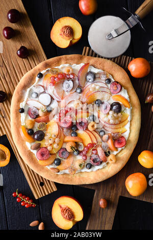 Gesunder Nachtisch. Flatlay Obst süße Pizza mit Pfirsichen, Aprikosen/Marillen, Kirschen, Johannisbeeren und Ricotta Creme auf dunklem Hintergrund overhead vie gemacht Stockfoto