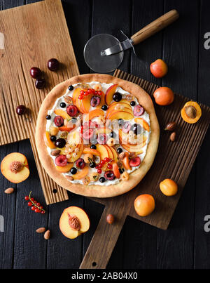 Gesundes Dessert flatlay. Obst Pizza mit raw Pfirsiche, Aprikosen, Kirschen, Johannisbeeren und Ricotta Creme auf dunklem Hintergrund Overhead Ansicht gemacht Stockfoto